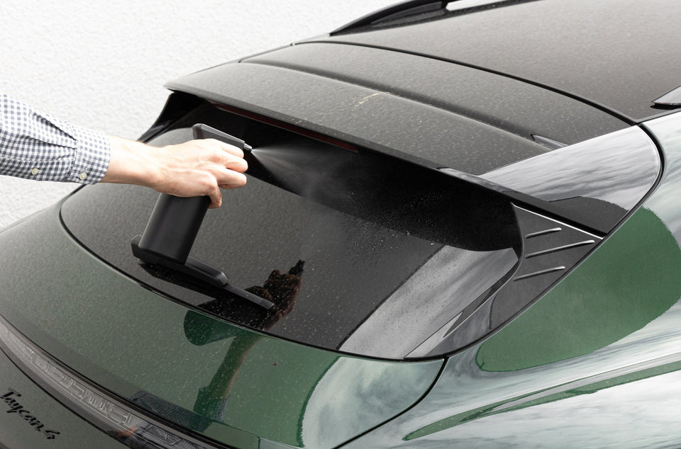 Hand sprüht den KULT Auto Glasreiniger auf die Heckscheibe eines dunkelgrünen Fahrzeugs