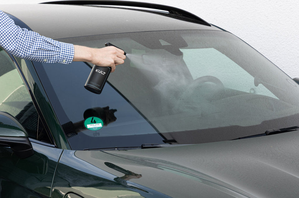 Hand sprüht den KULT Auto Glasreiniger aus der Sprühflasche auf die Windschutzscheibe eines dunkelgrünen Fahrzeugs