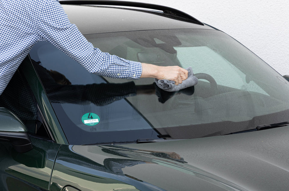 Hand wischt mit einem grauen Mikrofasertuch über die nasse Windschutzscheibe eines dunkelgrünen Fahrzeugs